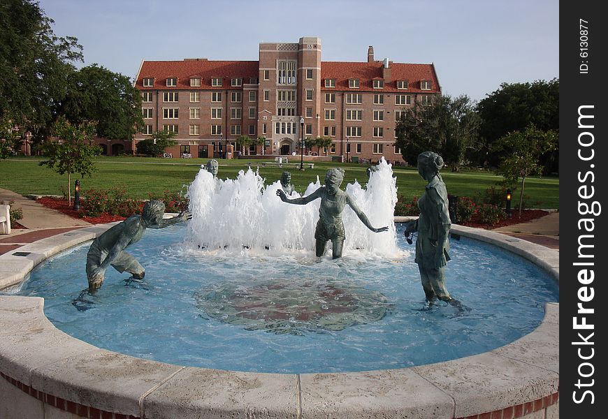 This is a water fountain that has bronze statues of people playing and having fun. There is a university dormitory in the background with blue skies.  This is a picture perfect college day.