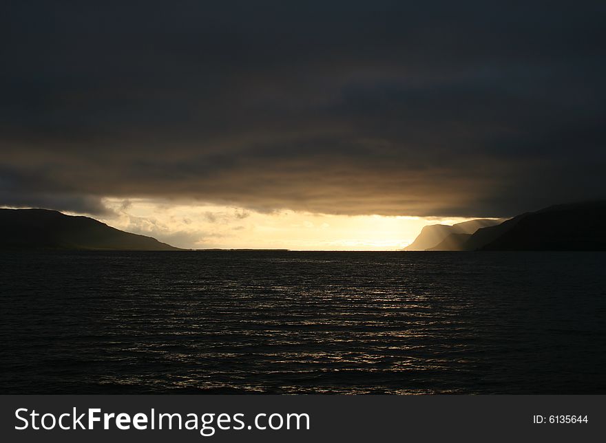 Golden cloudy sunset in a fjord with the clouds ending at the fjords mouth and the sun shining from behind them. Golden cloudy sunset in a fjord with the clouds ending at the fjords mouth and the sun shining from behind them.