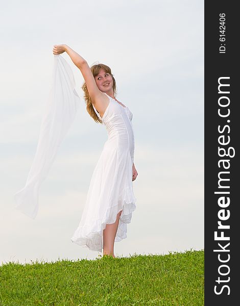 Girl with a white scarf in a hand stands on a green grass on a background of the blue sky