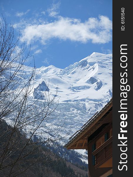 Snow slope of glacier against blue sky above mountain town, part of house and dark forest in the foreground. Snow slope of glacier against blue sky above mountain town, part of house and dark forest in the foreground