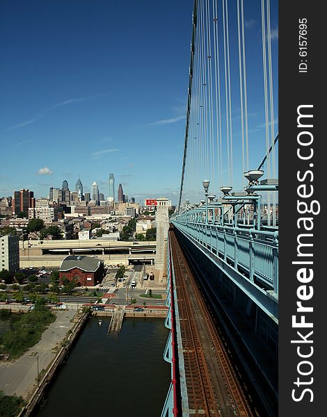 A view of a city from atop a bridge with blue railings, cables, and train tracks vanishing into the buildings, with the riverbank and docks below, on a beautiful sunny day against a clear blue sky. A shot of the new Philadelphia skyline from the Ben Franklin Bridge. A view of a city from atop a bridge with blue railings, cables, and train tracks vanishing into the buildings, with the riverbank and docks below, on a beautiful sunny day against a clear blue sky. A shot of the new Philadelphia skyline from the Ben Franklin Bridge.