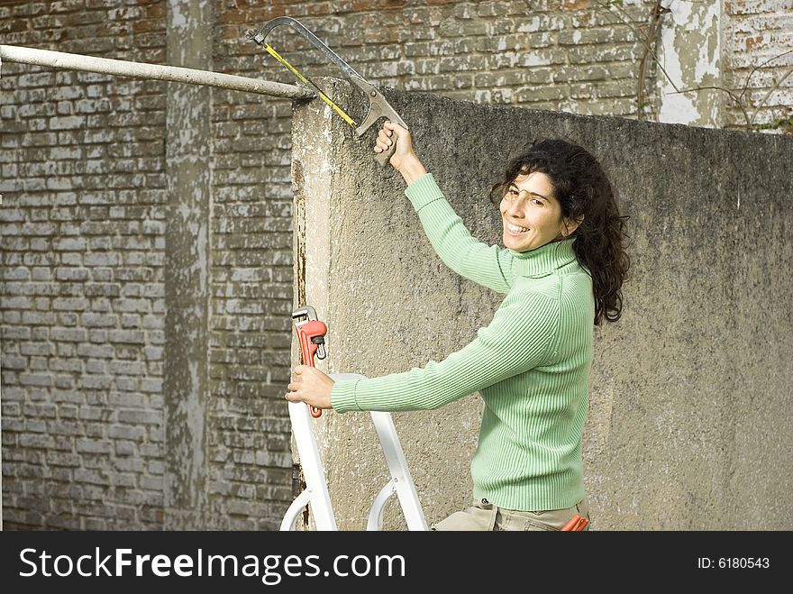 Woman stands on ladder while cutting pipe with hacksaw. She is smiling at camera. Vertically framed photo. Woman stands on ladder while cutting pipe with hacksaw. She is smiling at camera. Vertically framed photo.