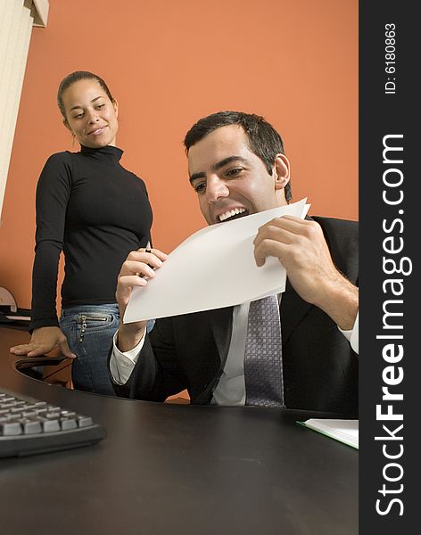 Businessman jokes around by putting a paper in his mouth as his co-worker watches. Vertically framed photo. Businessman jokes around by putting a paper in his mouth as his co-worker watches. Vertically framed photo