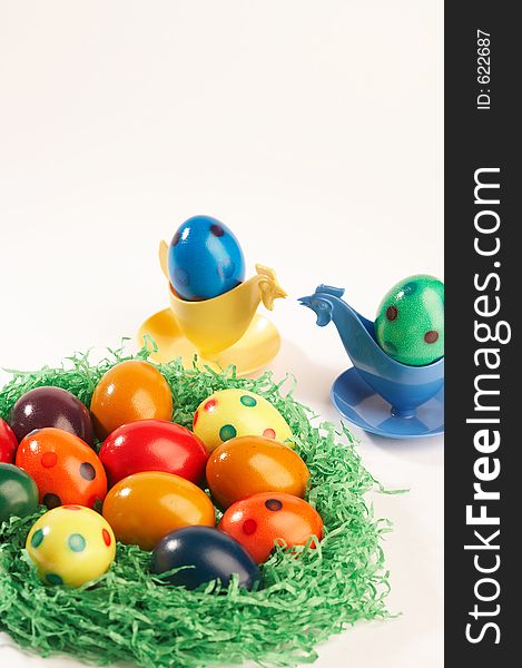 Coloured easter eggs in green grass with two eggcups looking like hens on white background