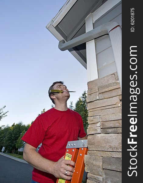 Man with a  screwdriver in his mouth standing on a ladder looking up at a house. Vertically framed photo. Man with a  screwdriver in his mouth standing on a ladder looking up at a house. Vertically framed photo.