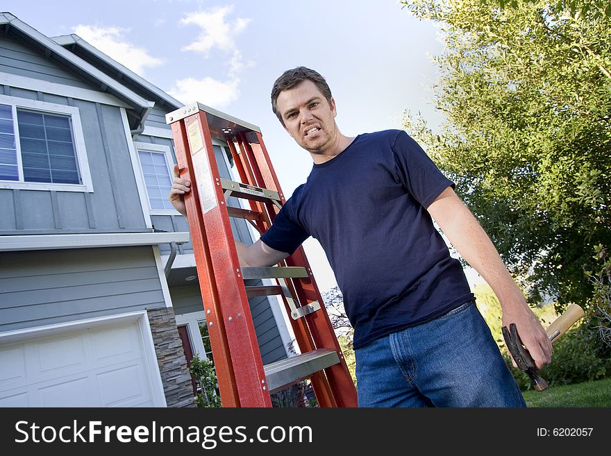 Unhappy man standing in front of house holding ladder and hammer. Horizontally framed photo. Unhappy man standing in front of house holding ladder and hammer. Horizontally framed photo.