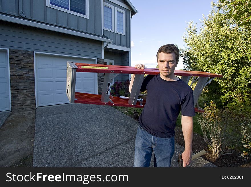 Frowning man standing in front of house holding ladder and hammer. Horizontally framed photo. Frowning man standing in front of house holding ladder and hammer. Horizontally framed photo.