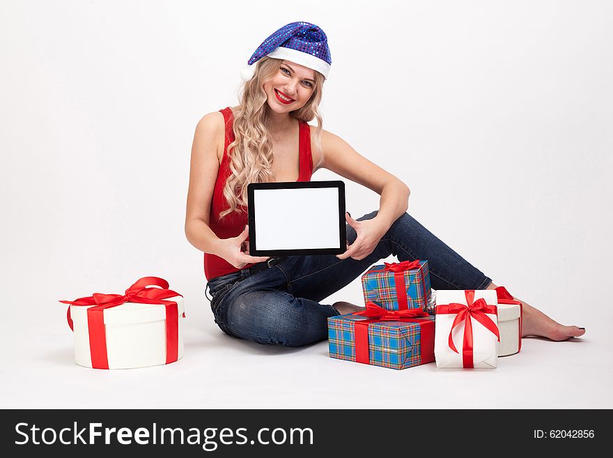 The beautiful blonde with big breasts is sitting on a white background and holds a a tablet in blue New Year's cap and smiling at the camera, selective focus on the tablet, picture with depth of field