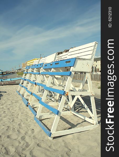 A bright daylight shot of a row of blue and white painted wooden lifeguard chairs on a sandy beach, with lifeboats, a beach boardwalk, and a clear blue sky behind them. A bright daylight shot of a row of blue and white painted wooden lifeguard chairs on a sandy beach, with lifeboats, a beach boardwalk, and a clear blue sky behind them