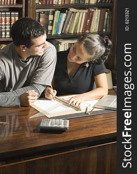 Two students smiling at each other as they work at a desk with a laptop, calculator, ruler, and calculator on it. Vertically framed photo. Two students smiling at each other as they work at a desk with a laptop, calculator, ruler, and calculator on it. Vertically framed photo.