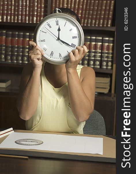 Woman at a desk holding a clock up in front of her face. Vertically framed photograph. Woman at a desk holding a clock up in front of her face. Vertically framed photograph.