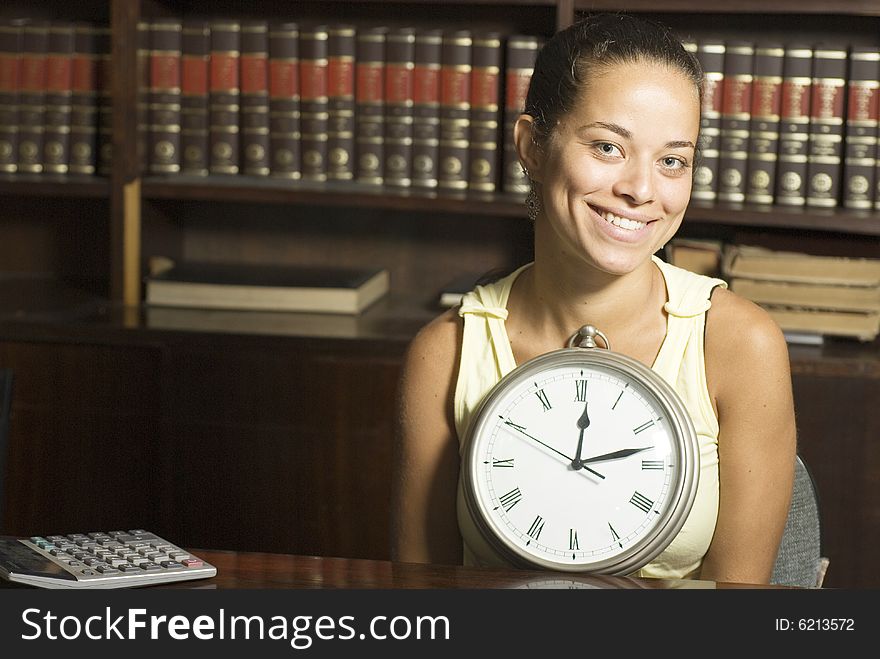 Smiling student holding a clock while seated at a desk in an office surrounded by books. Horizontally framed photo. Smiling student holding a clock while seated at a desk in an office surrounded by books. Horizontally framed photo.