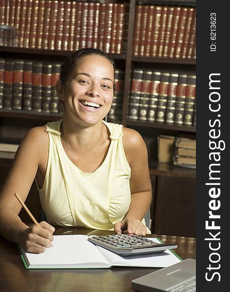 Happy student writing in a notebook with a pencil, and a laptop on her desk and books in the background. Vertically framed photo. Happy student writing in a notebook with a pencil, and a laptop on her desk and books in the background. Vertically framed photo.