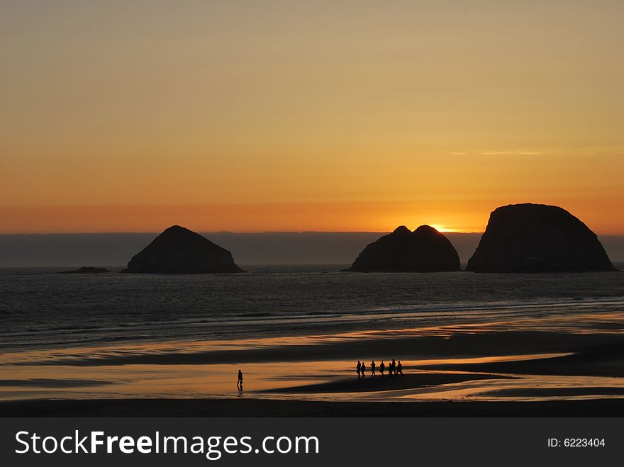 A group of young people take a walk on the beach as the sun sets behind three rocks in the ocean. A group of young people take a walk on the beach as the sun sets behind three rocks in the ocean.