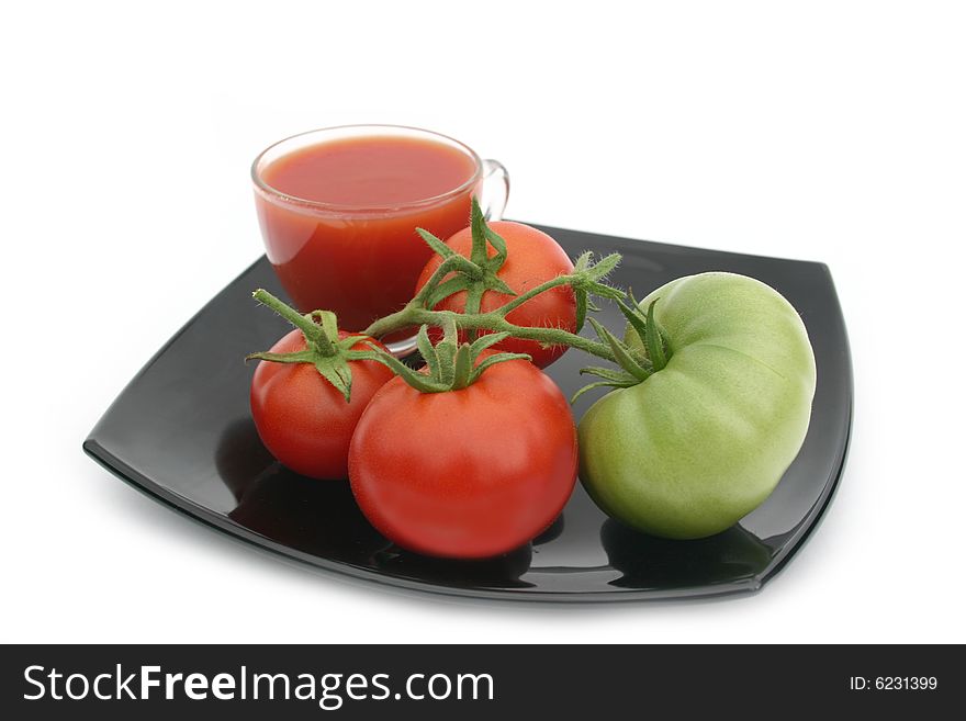 Red tomatoes and a green tomato on a black plate with a transparent cup of tomato juice on a white background
