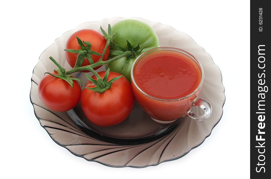 Red tomatoes and a green tomato on a plate with a transparent cup of tomato juice on a white background