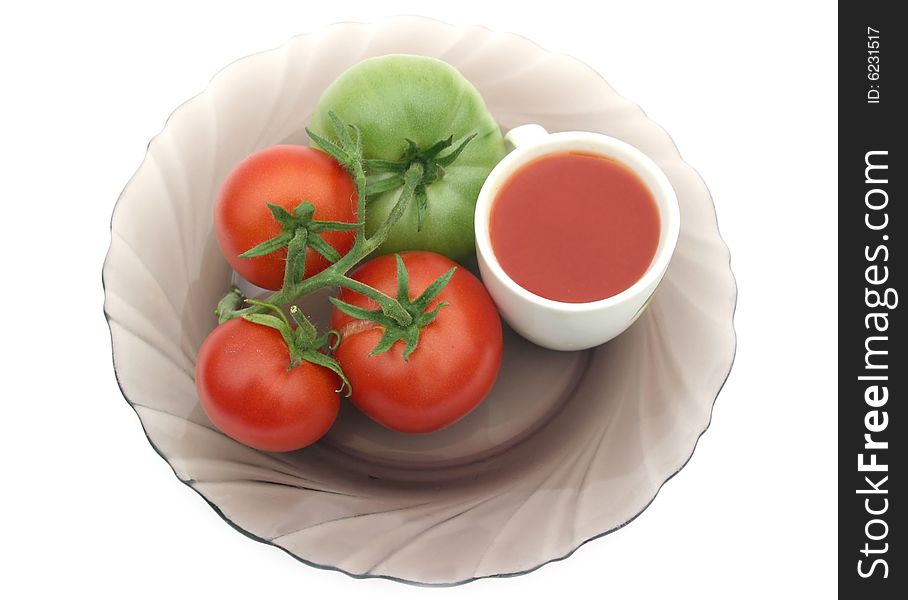 Red tomatoes and a green tomato on a plate with a cup of tomato juice on a white background