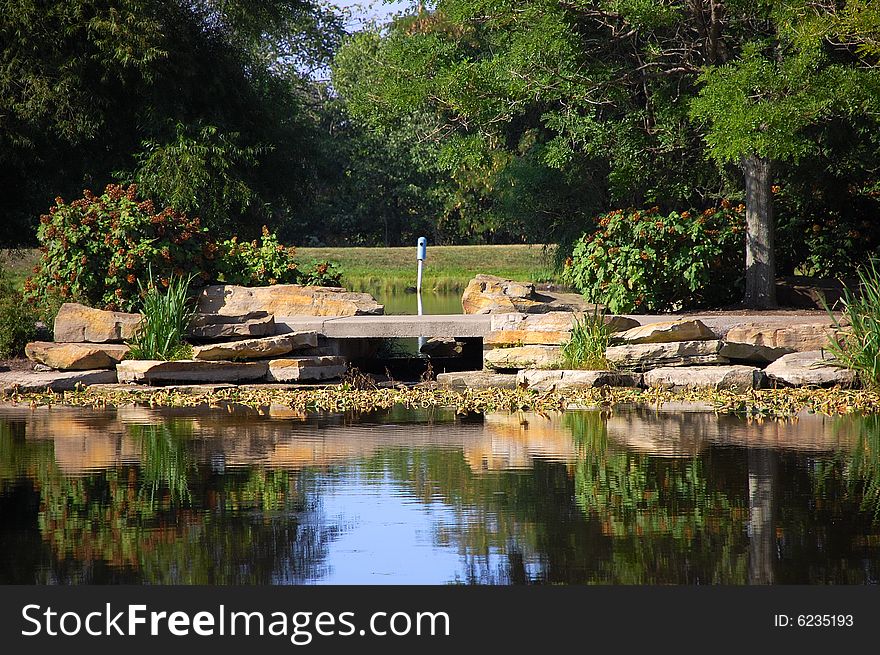 This is a slab stone bridge. The shot was taken from across  the lake, which is in the foreground with reflections. Trees shade the background.