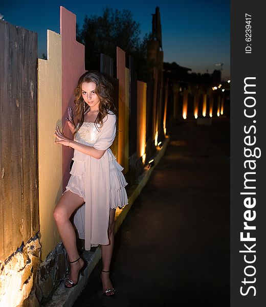 Girl teenager stands outdoor on evening in illumination near color fence against a night sky background. Girl teenager stands outdoor on evening in illumination near color fence against a night sky background.