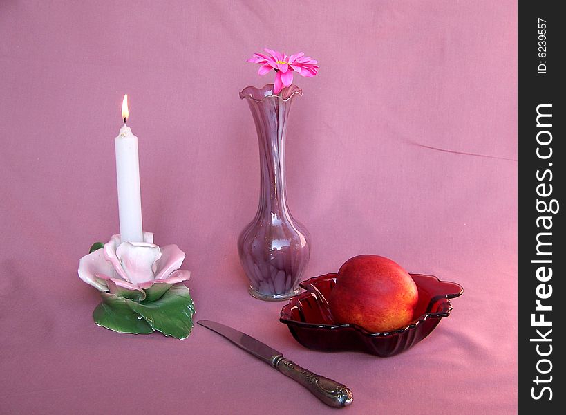 Candle in candlestick, vase with flower, peach and knife on the pink background. Candle in candlestick, vase with flower, peach and knife on the pink background