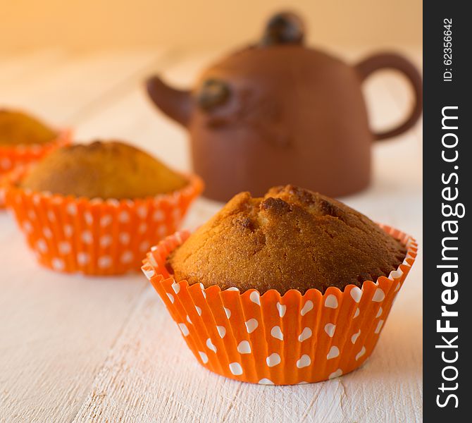 Muffins in orange paper cupcake holder with white polka dots and brown ceramic teapot on a white wooden background, selective focus, square. Muffins in orange paper cupcake holder with white polka dots and brown ceramic teapot on a white wooden background, selective focus, square