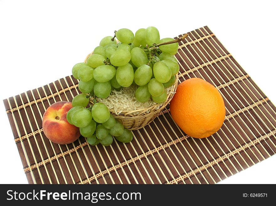 Green grapes with an orange and a 
peach at a yellow basket on a white background