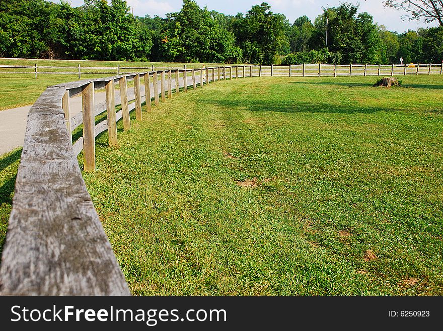 This is a shot of a wood fence used to keep horses off the road. The road can be seen in the shot. Green is a strong color in the image, the sky can also be seen. This is a shot of a wood fence used to keep horses off the road. The road can be seen in the shot. Green is a strong color in the image, the sky can also be seen.