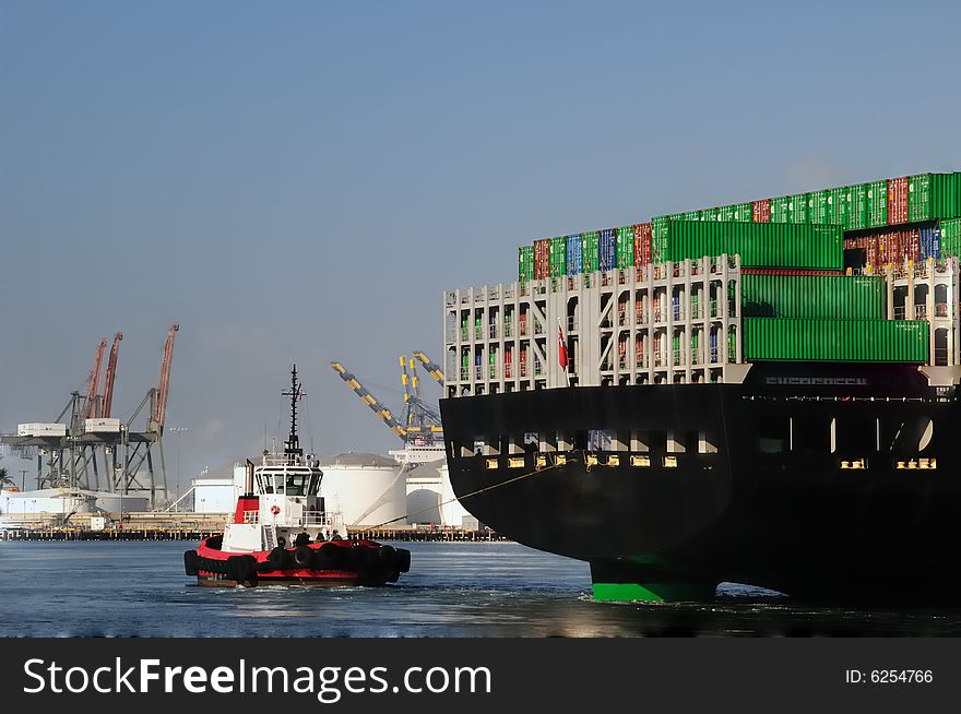 View from behind as a red tugboat pulls a massive merchant vessel around in a narrow harbor channel, with loading cranes in the background. View from behind as a red tugboat pulls a massive merchant vessel around in a narrow harbor channel, with loading cranes in the background.