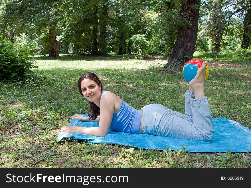 A girl, lying on a blue towel in the middle of the park, doing some exercises with a ball. A girl, lying on a blue towel in the middle of the park, doing some exercises with a ball