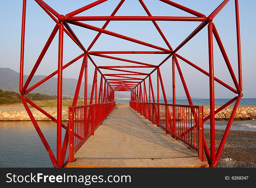 Red cantilever bridge of structural steel with wooden floor, over the mouth of the creek near the Aegean sea in Gumuldur, Turkey. A bicycle parked at the far end. Rocks form the bank of the river. Red cantilever bridge of structural steel with wooden floor, over the mouth of the creek near the Aegean sea in Gumuldur, Turkey. A bicycle parked at the far end. Rocks form the bank of the river.