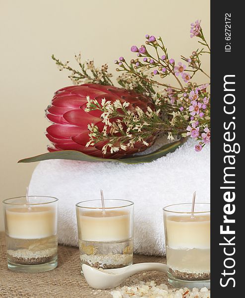 Relaxing spa scene with a white rolled up towel, protea and pink flowers, beautiful handmade candles and bath salts