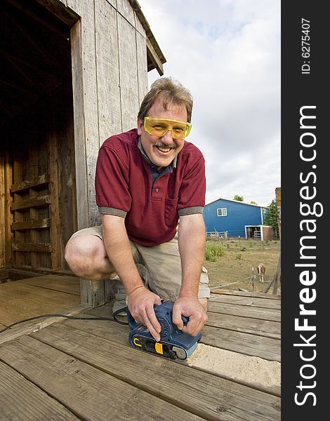 Smiling man with safety glasses using a power sander on some wood. Vertically framed photo. Smiling man with safety glasses using a power sander on some wood. Vertically framed photo.