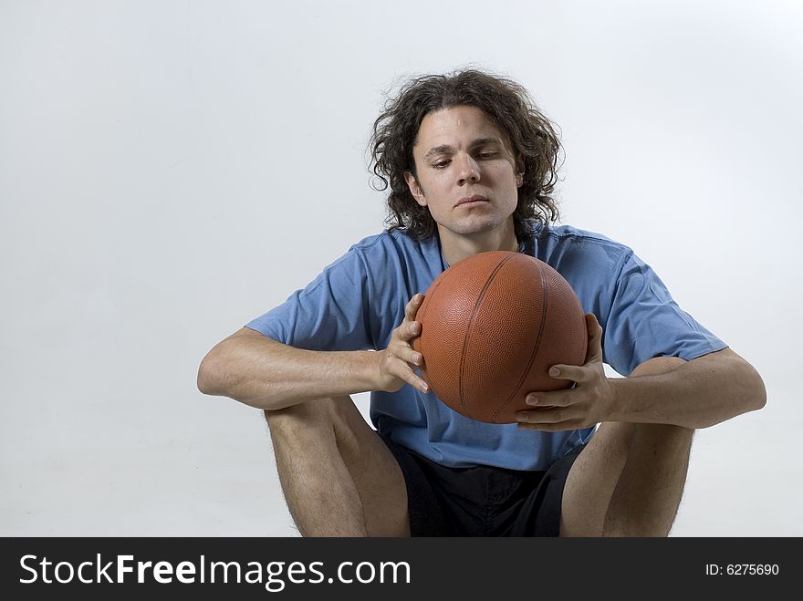 A young man sitting. He is holding a basketball in his hands. His eyes are focused on the ball and he has a serious expression on his face. Horizontally framed shot. A young man sitting. He is holding a basketball in his hands. His eyes are focused on the ball and he has a serious expression on his face. Horizontally framed shot.