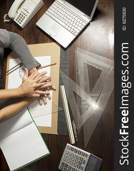 Two hands on desk surrounded by drafting tools, laptop, phone, and calculator. Vertically framed photo.