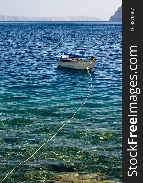 A lonely colorful small boat floating in the crystal clear Aegian Sea, Greece, near Thirassia. In the background Thira (Santorini) emerges from the sea. A lonely colorful small boat floating in the crystal clear Aegian Sea, Greece, near Thirassia. In the background Thira (Santorini) emerges from the sea.