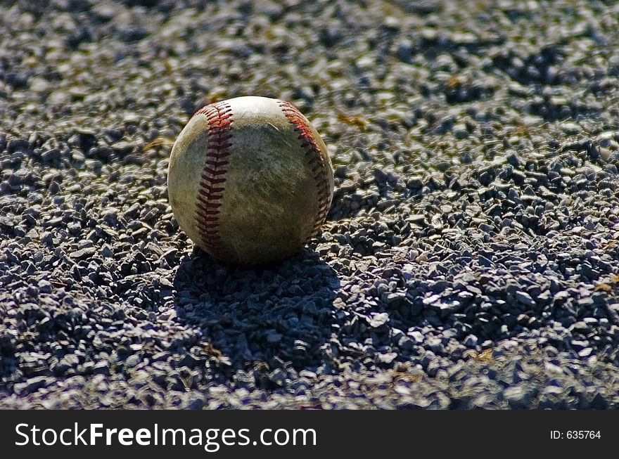 A well-used baseball sits in the gravel at the side of the field casting a shadow from the afternoon sun. A well-used baseball sits in the gravel at the side of the field casting a shadow from the afternoon sun.