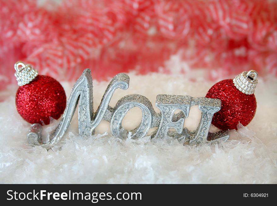 The word Noel made of glitter sitting in the middle of fake snow with two red christmas ornaments on each end.  Used selective focus and a shallow depth of field. The word Noel made of glitter sitting in the middle of fake snow with two red christmas ornaments on each end.  Used selective focus and a shallow depth of field.