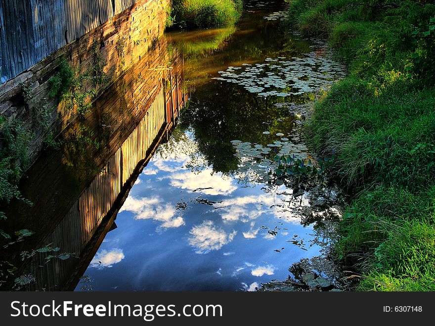 The side of an old barn and sky are reflected in a quiet summer stream. The side of an old barn and sky are reflected in a quiet summer stream.
