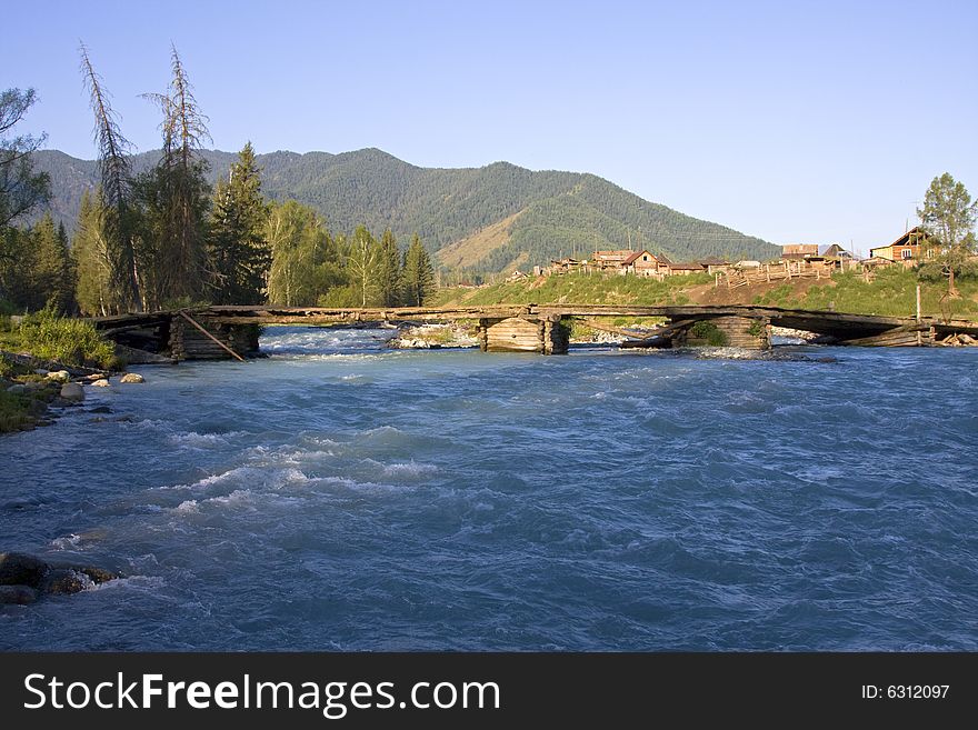 Village and the wooden bridge through a wide rapid mountain river in forest, Altai, Russia. Village and the wooden bridge through a wide rapid mountain river in forest, Altai, Russia