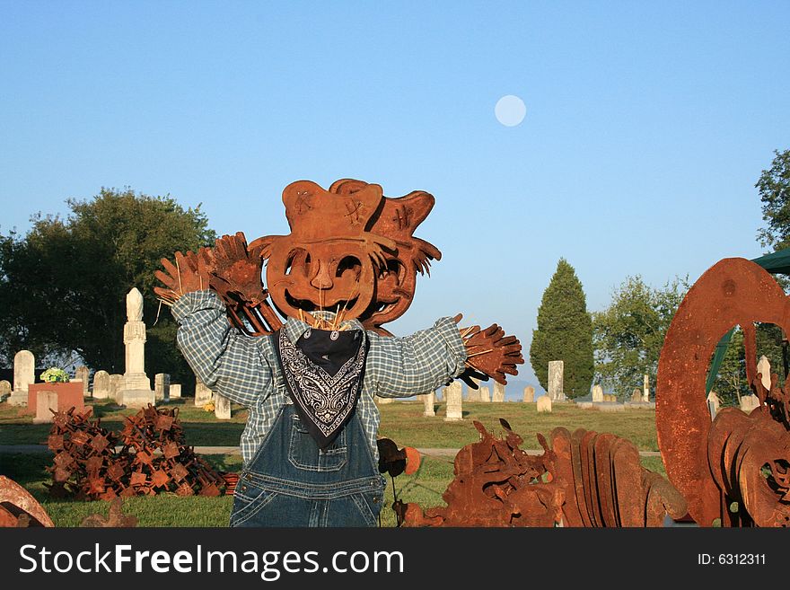 Metal pumpkin scarecrow dressed in overalls with an old time cemetery in the back ground with a early morning cloudless blue sky with a full moon
