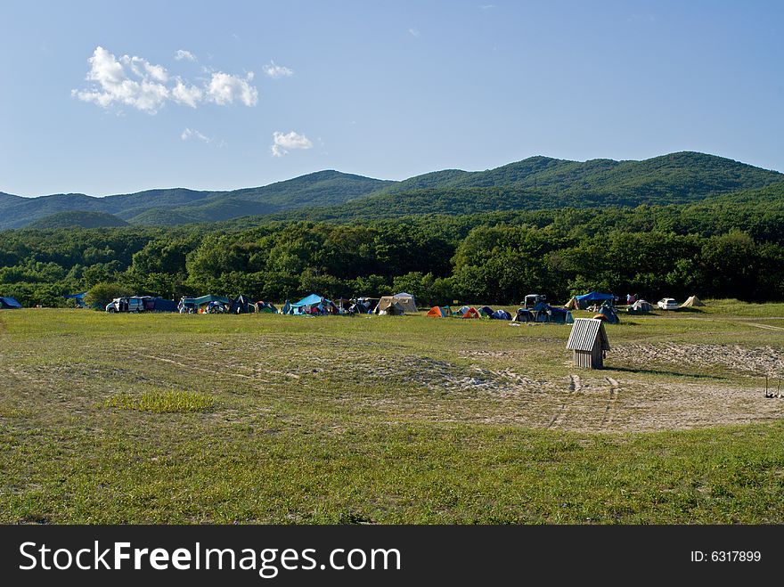 A camp of touristes on broad meadow at seacoast. On background are green hills and blue sky with clouds. A camp of touristes on broad meadow at seacoast. On background are green hills and blue sky with clouds.