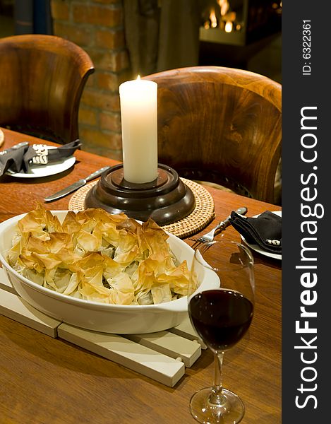 A romantic table setting with candle and fireplace in the background, with a gourmet chicken pie and glass of red wine on a beautiful wooden table.
