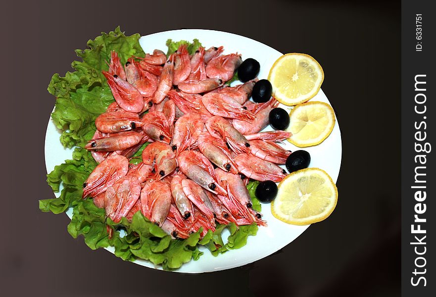 Dish with boiled shrimps decorated with olives, salad and segments of lemon on the black background. Dish with boiled shrimps decorated with olives, salad and segments of lemon on the black background