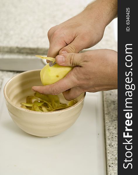 Hands peeling potatoes into a bowl on a counter next to a bowl and tomatoes. Vertically framed photo.