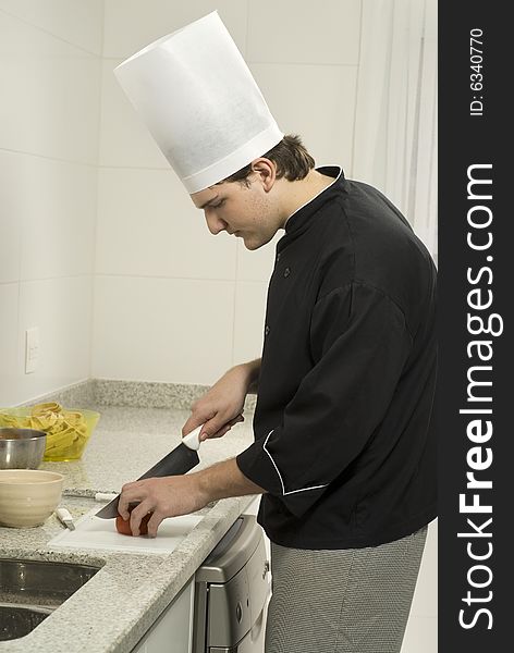 Young male chef in a kitchen slicing tomatoes in a bowl with a large knife. Vertically framed photo.