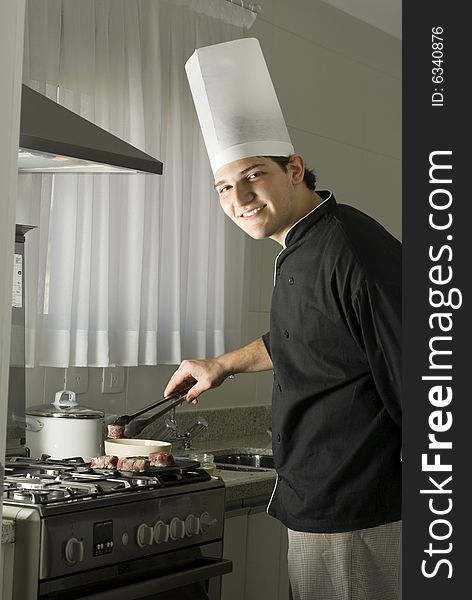 Young, smiling chef grilling steaks on a griddle on a stove in a kitchen. Vertically framed photo.