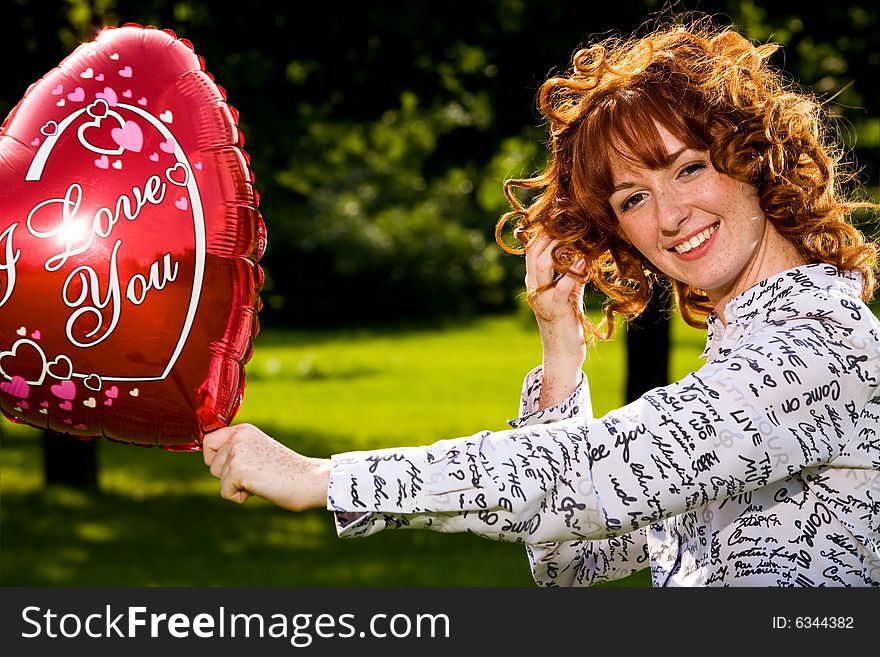 Red-haired lovely young woman holding red balloon in shape of heart outdoors. Red-haired lovely young woman holding red balloon in shape of heart outdoors