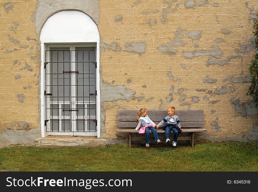 A four year-old girl and boy sit on a bench holding hands in front of a wall of an old, non-descript building. A four year-old girl and boy sit on a bench holding hands in front of a wall of an old, non-descript building.