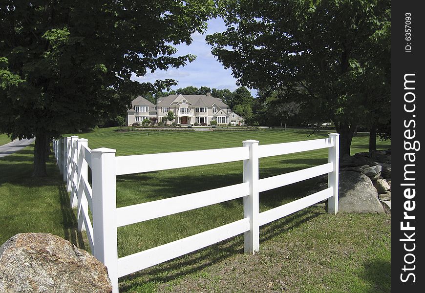 View of large, white house with lawns, gardens and white fence, framed with trees and blue sky. View of large, white house with lawns, gardens and white fence, framed with trees and blue sky