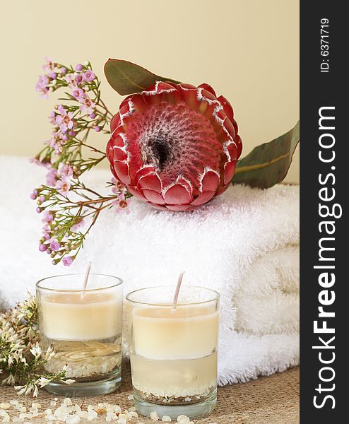 Relaxing spa scene with a white rolled up towel, pink flowers and protea, beautiful handmade candles and bath salts
